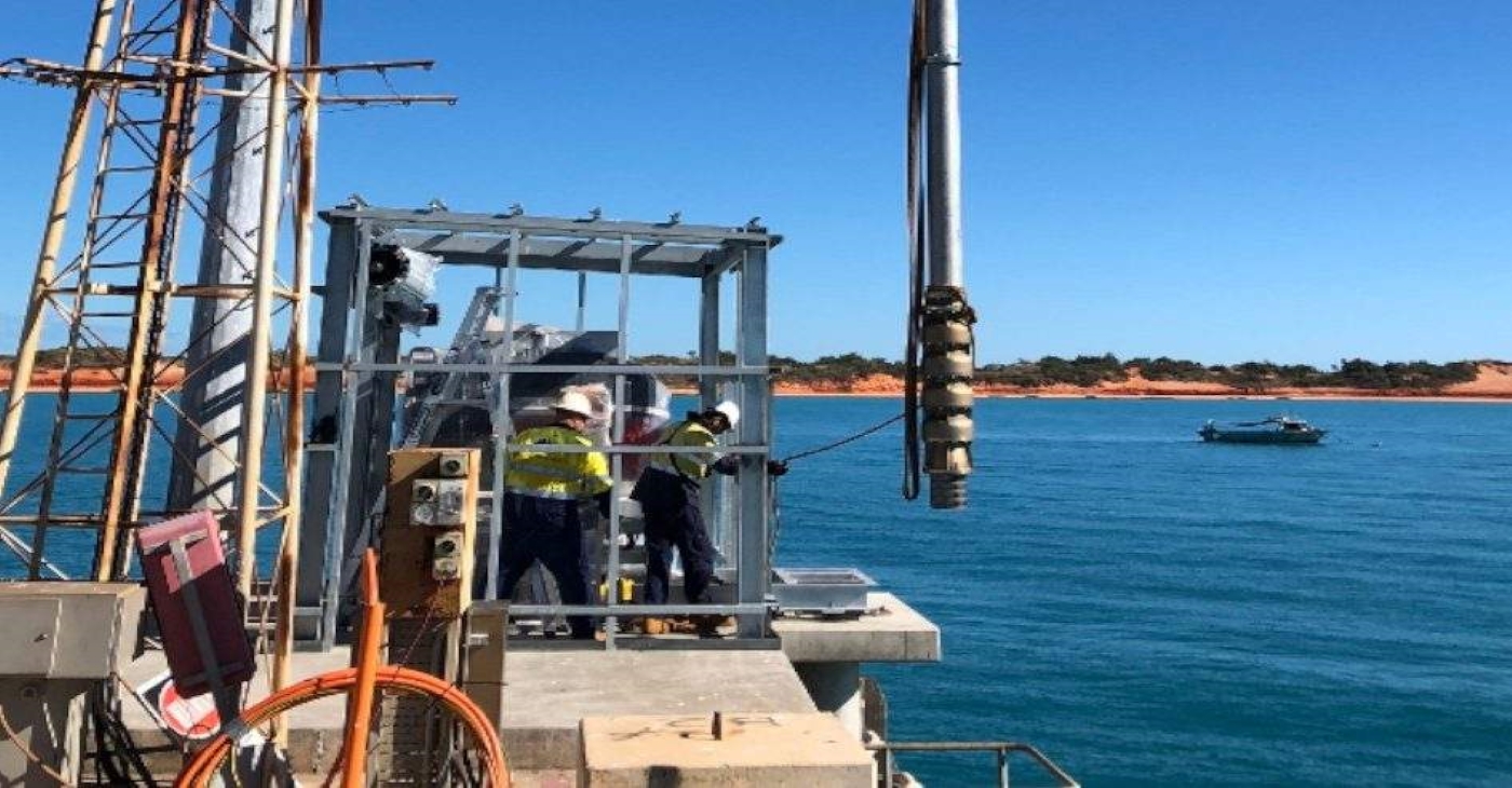 Industrial crew installing a large vertical turbine fire pump from a jetty structure over the ocean, illustrating a marine or offshore fire suppression system installation