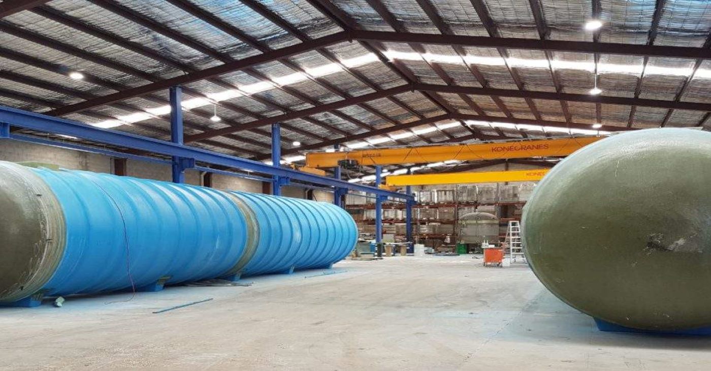 Manufacturing facility interior showing large blue and green FRP (Fiberglass Reinforced Plastic) pump station vessels in production, used for municipal and industrial wastewater handling