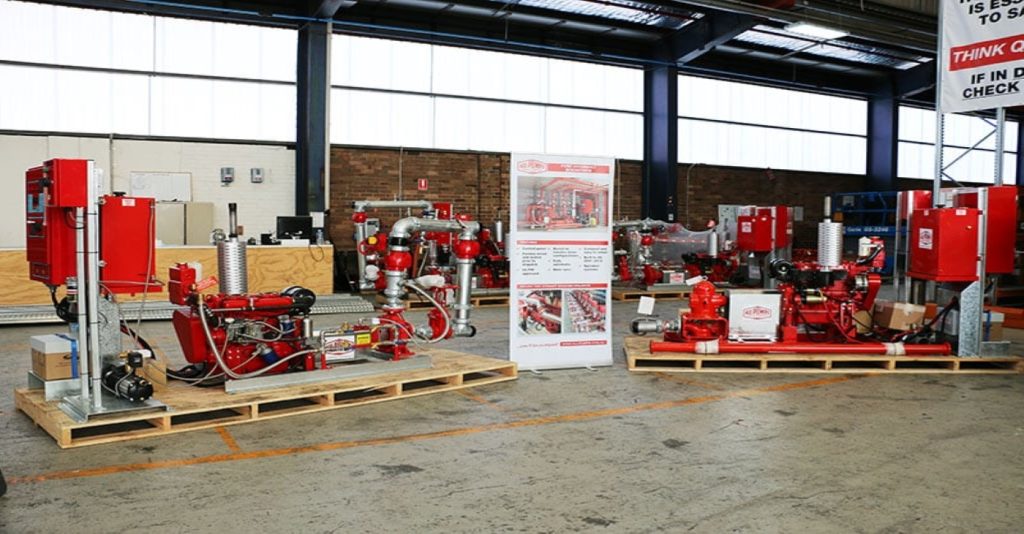 Multiple red fire pump systems on wooden pallets are arranged in a warehouse, with a standing banner displaying product specs.