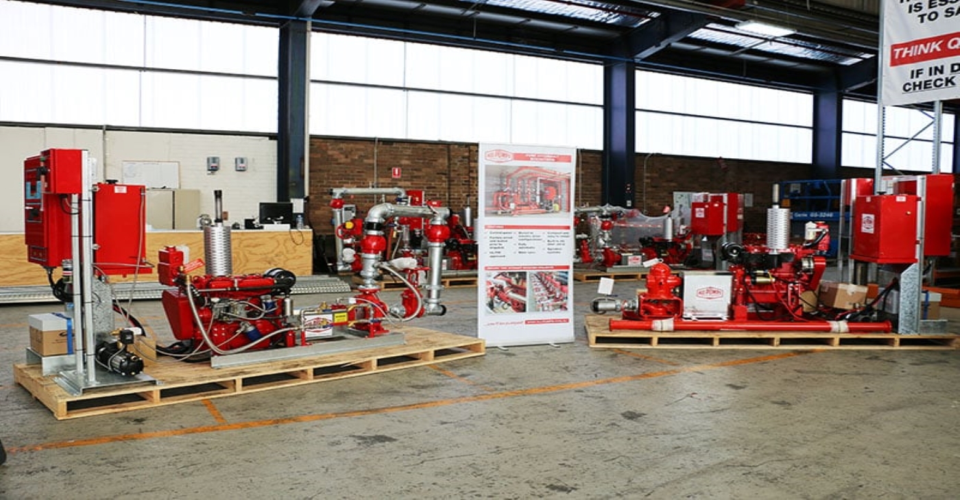Multiple red fire pump systems on wooden pallets are arranged in a warehouse, with a standing banner displaying product specs.