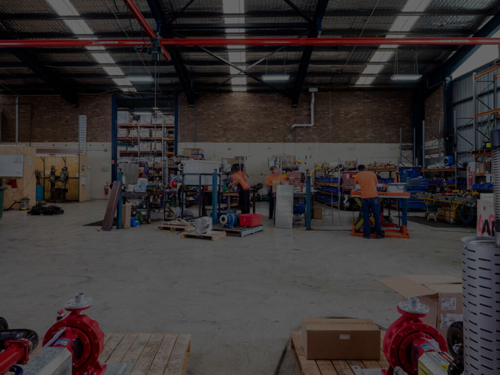 Wide view of the industrial ALL-PUMPS warehouse and workshop floor, showing technicians working on pump assembly and repair, with various pump components and red centrifugal pumps visible in the foreground