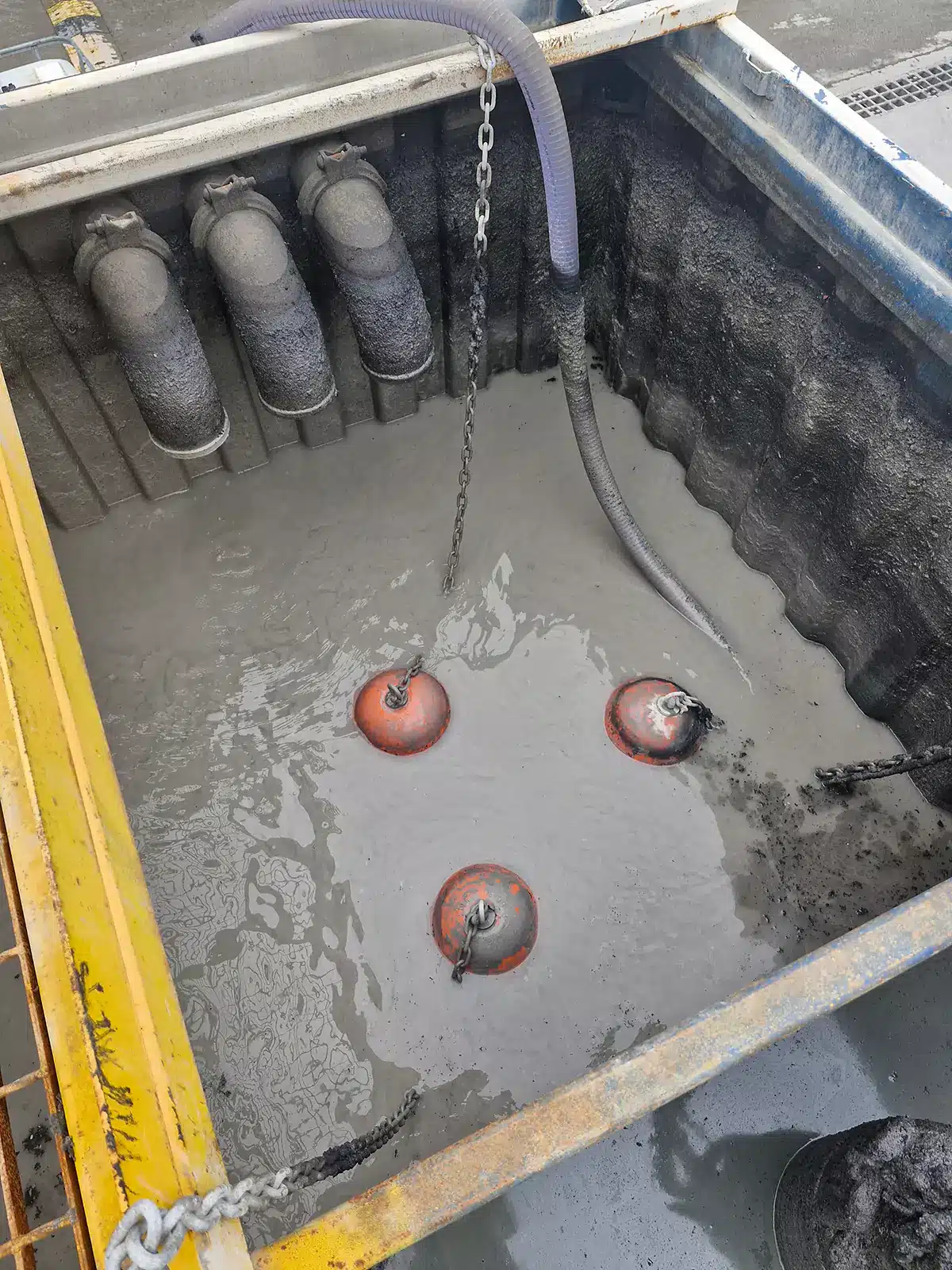 Industrial pit filled with thick sludge and oil-water mixture, showing a hose and three anchor points for an oil skimmer system used in wastewater treatment and contamination control